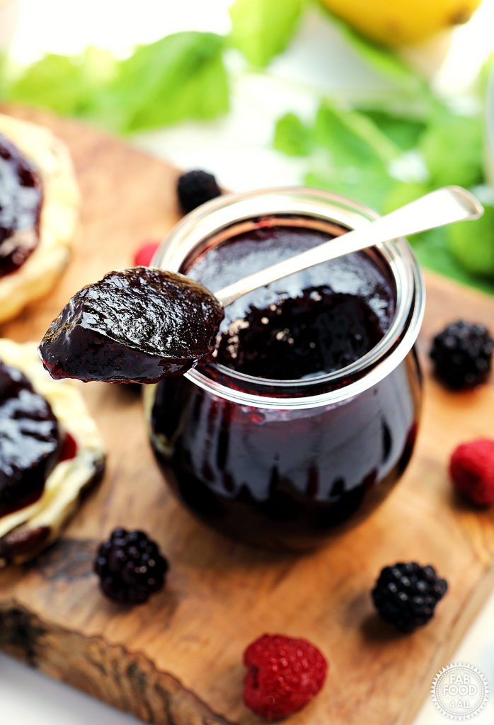 A teaspoon of Seedless Blackberry & Raspberry jam resting on top of a jar on a board surrounded by berries, 2 scone halves, raspberry leaves and a lemon.