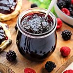 Pot of Seedless Blackberry & Raspberry Jam with a spoon in it on a board with scones, a bowl of berries and some raspberry leaves in the background.