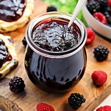 Pot of Seedless Blackberry & Raspberry Jam with a spoon in it on a board with scones, a bowl of berries and some raspberry leaves in the background.