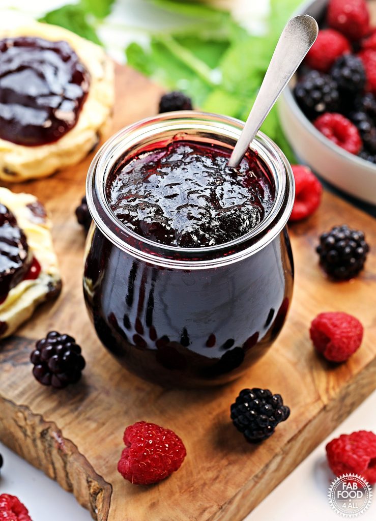 Pot of Seedless Blackberry & Raspberry Jam with a spoon in it on a board with scones, a bowl of berries and some raspberry leaves in the background.