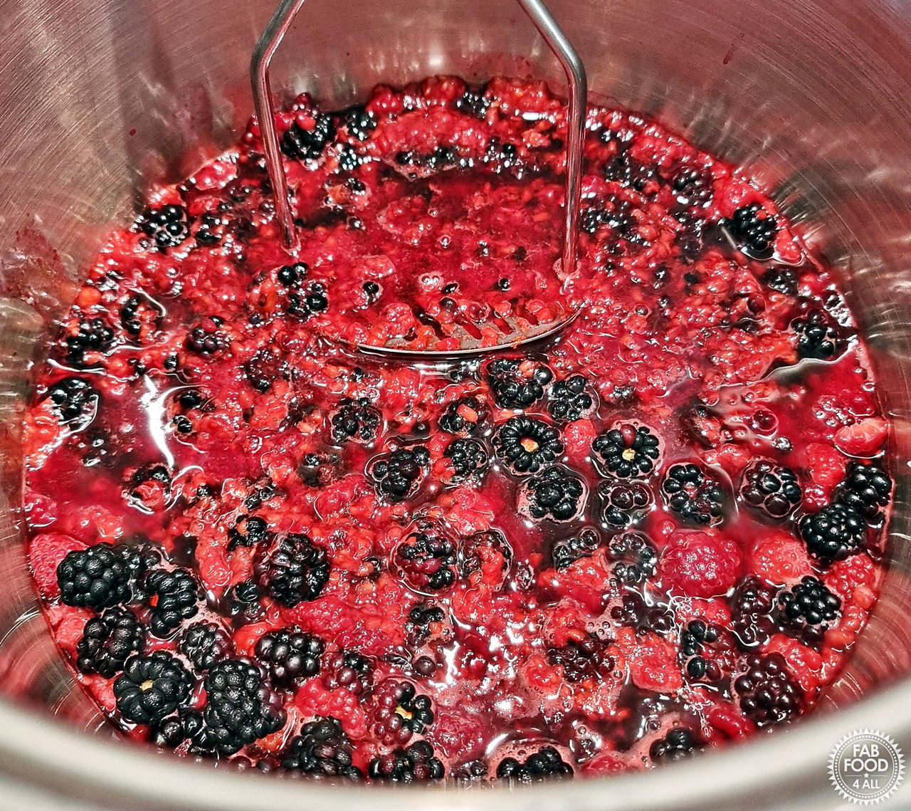 Blackberries and raspberries being crushed with a potato masher in a large stainless steel pot as they're brought to the boil.