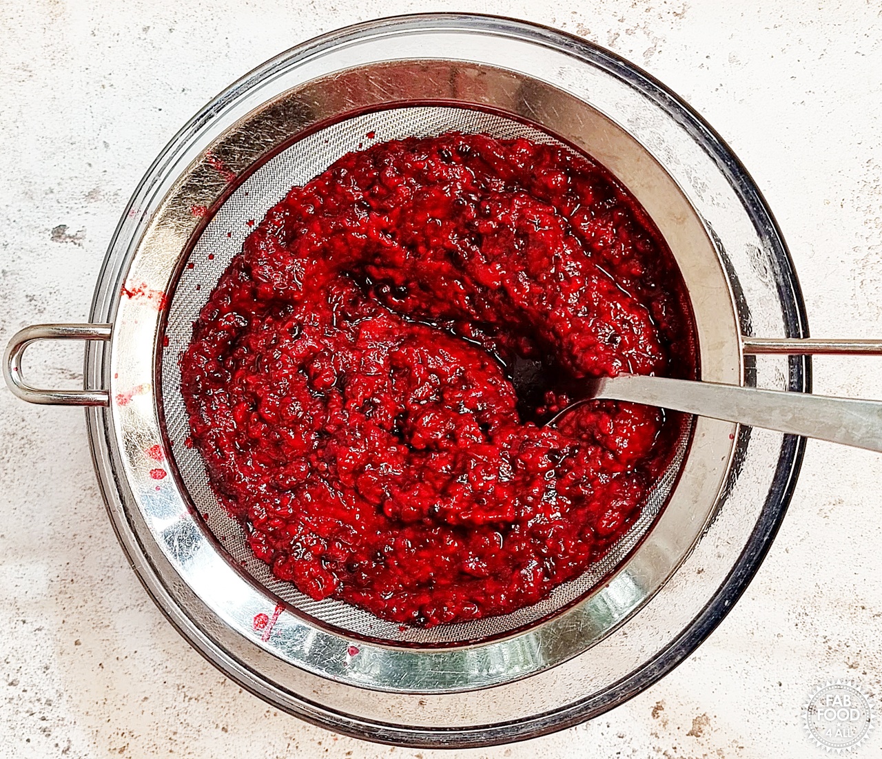 Cooked blackberries and raspberries in a sieve over a glass bowl with large metal spoon pushing the pulp.