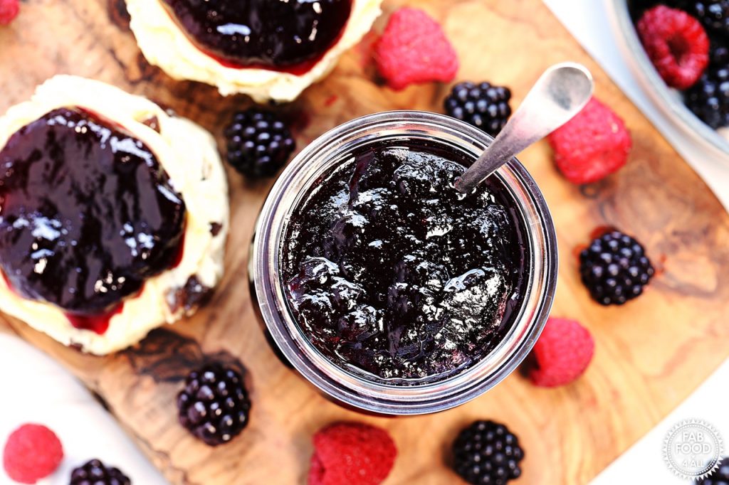 Overhead shot of open jar of Seedless Blackberry & Raspberry Jam with a teaspoon in it on a board with scones and berries.
