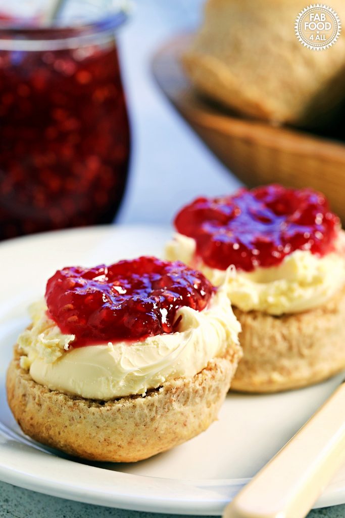 2 Spelt scones halves on a plate with clotted cream and raspberry jam. Pot of jam and bowl or scones just visible in the background.