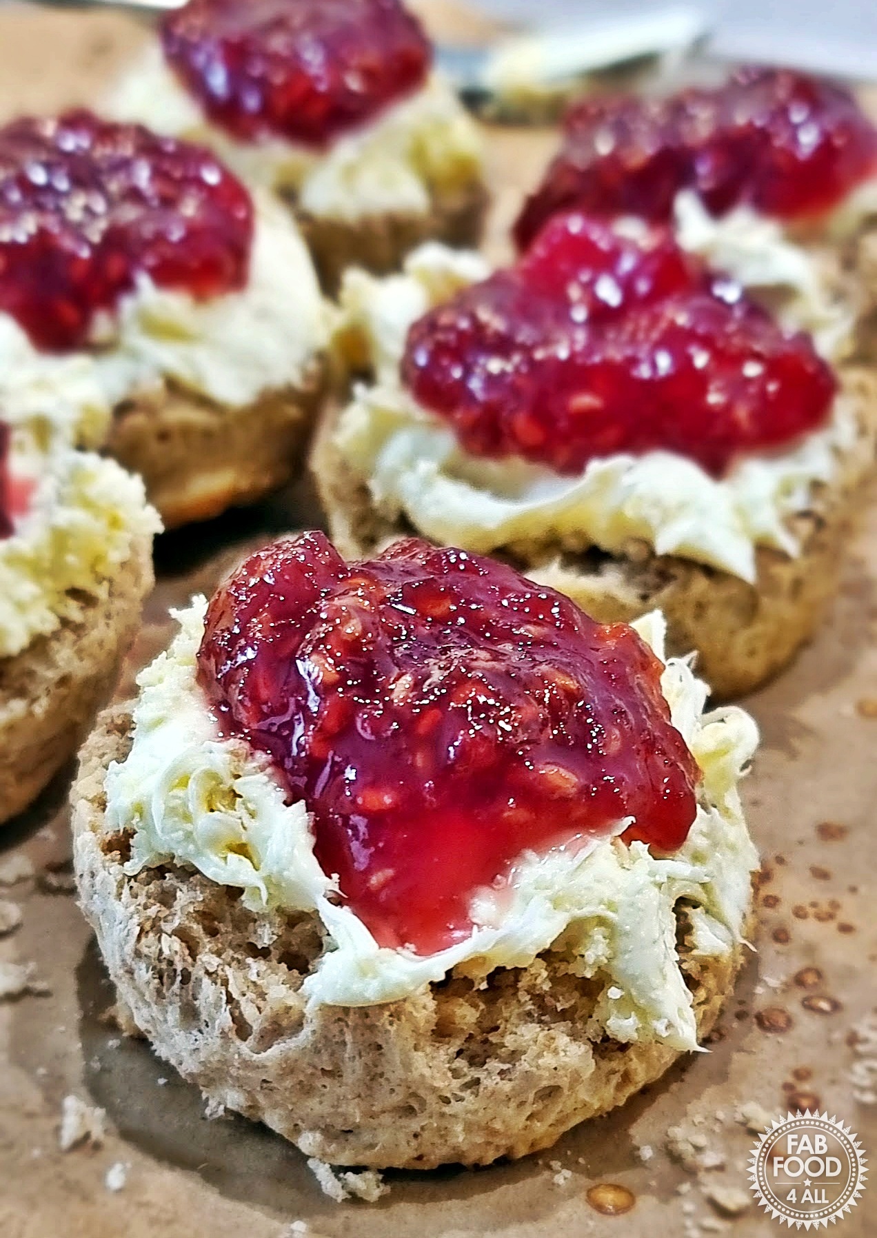 Spelt scones with clotted cream and raspberry jam on parchment with teaspoon in the background.