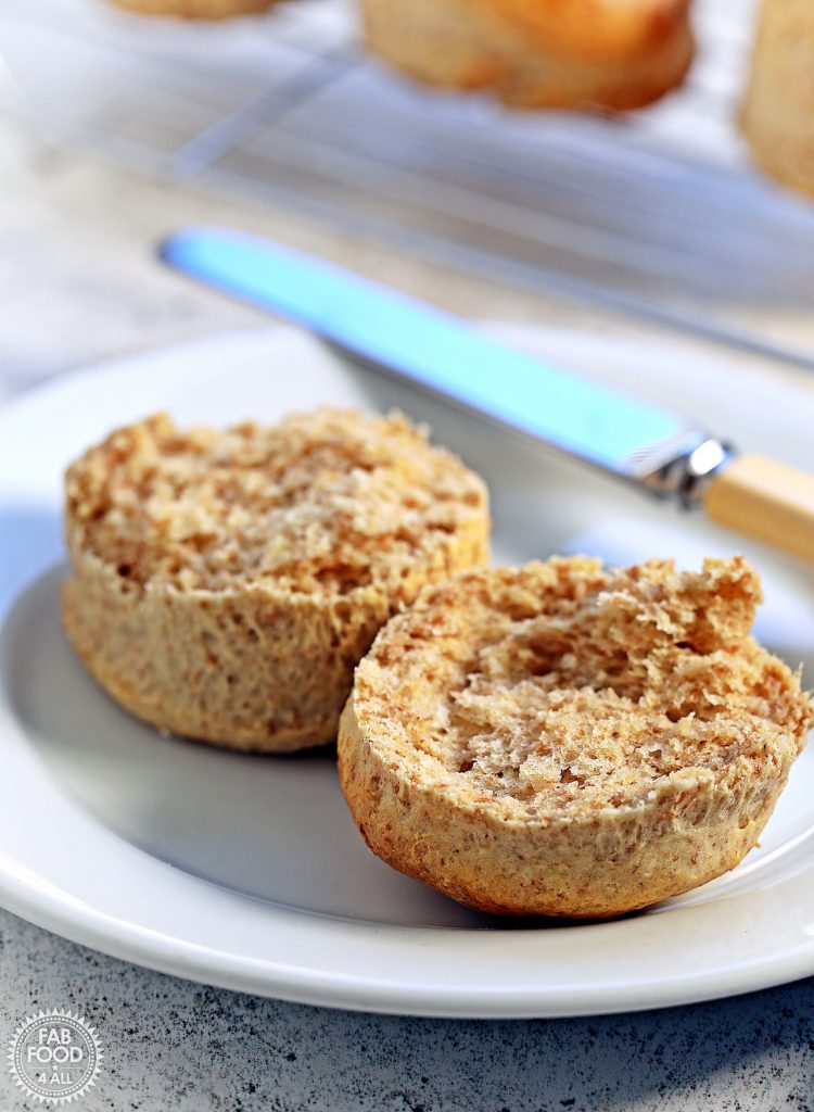A spelt scone split into 2 on a plate with a butter knife and wire rack with more scones just visible in the background.