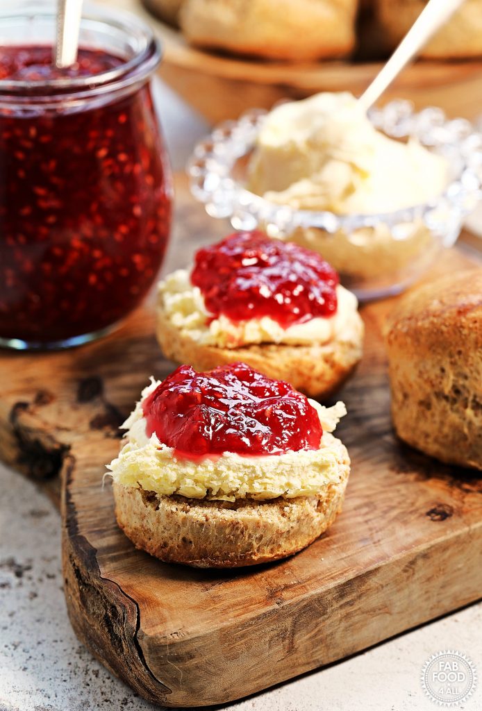 Spelt Scones on a wooden board with clotted cream and raspberry jam. Bowl of scones just visible in the background.