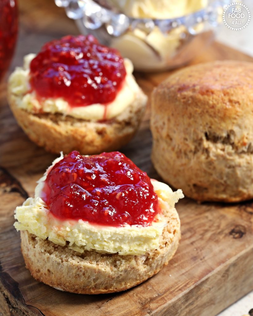 Close up of spelt scones with clotted cream and raspberry jam (2 halves) on a wooden block with whole scone and glass dish of clotted cream in background.