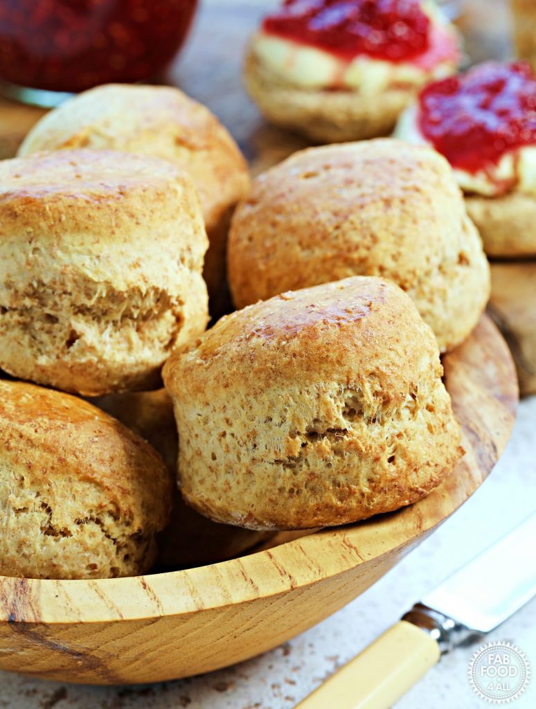 Wooden bowl of spelt scones with butter knife just visible and scones with jam and clotted cream in the background with a pot of jam.