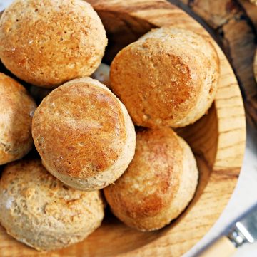 Overhead shot of a wooden bowl of spelt scones with butter knife and edge of a scone with jam and clotted cream just visible in corners.
