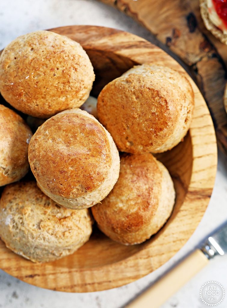Overhead shot of a wooden bowl of spelt scones with butter knife and edge of a scone with jam and clotted cream just visible in corners.