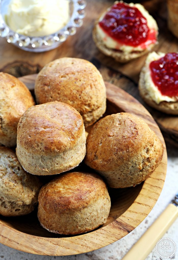 Spelt Scones in a wooden bowl with dish of clotted cream and 2 scones halves with raspberry jam and clotted cream in the background on a wooden block.