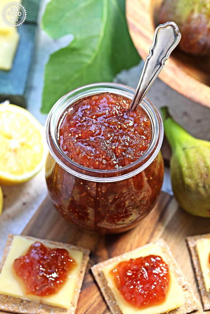 Simple Fig Jam on a wooden board with a teaspoon resting in it with cheese & biscuits topped with fig jam in the foreground. Fig leaf, halved lemon, bowl of figs and marble cheese block just visible in background.