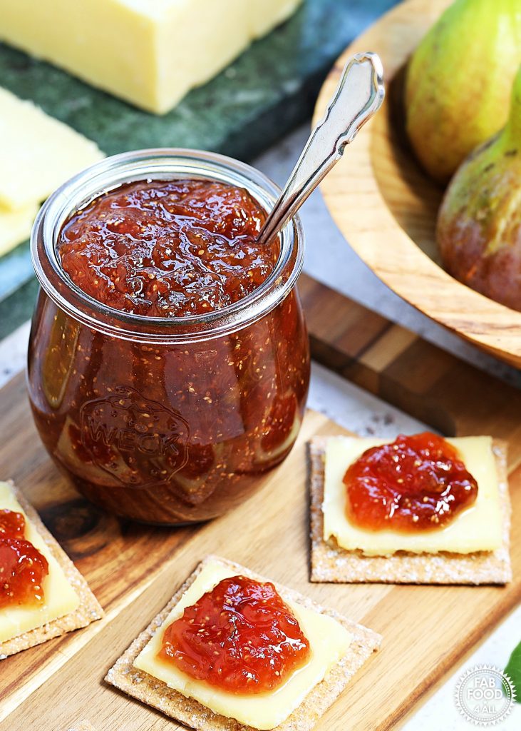 Simple Fig Jam in a glass jar with teaspoon on a wooden board with cheese and crackers topped with fig jam. Bowl of figs and block of Cheddar in the background.