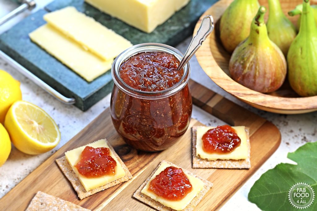Simple Fig Jam in a glass jar with teaspoon on a wooden board with cheese and crackers topped with fig jam. Bowl of figs, halved lemons and block of Cheddar in the background.