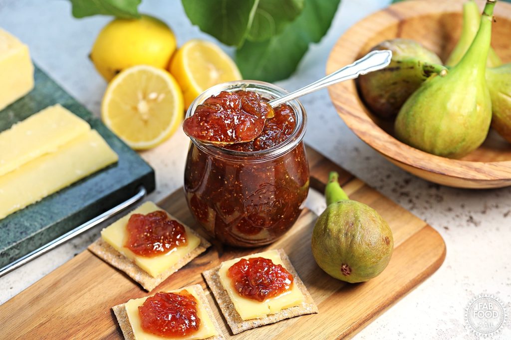Simple Fig Jam in a glass jar with teaspoon resting on top of it on a wooden board with cheese and crackers topped with fig jam. Bowl of figs, halved lemons, fig leaves and Cheddar on a marble board in the background.