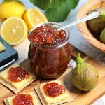Simple Fig Jam in a glass jar with teaspoon resting on top of it on a wooden board with cheese and crackers topped with fig jam. Bowl of figs, halved lemons, fig leaves and Cheddar on a marble board just visible.