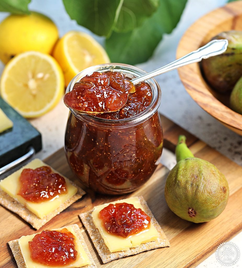 Simple Fig Jam in a glass jar with teaspoon resting on top of it on a wooden board with cheese and crackers topped with fig jam. Bowl of figs, halved lemons, fig leaves and Cheddar on a marble board just visible.
