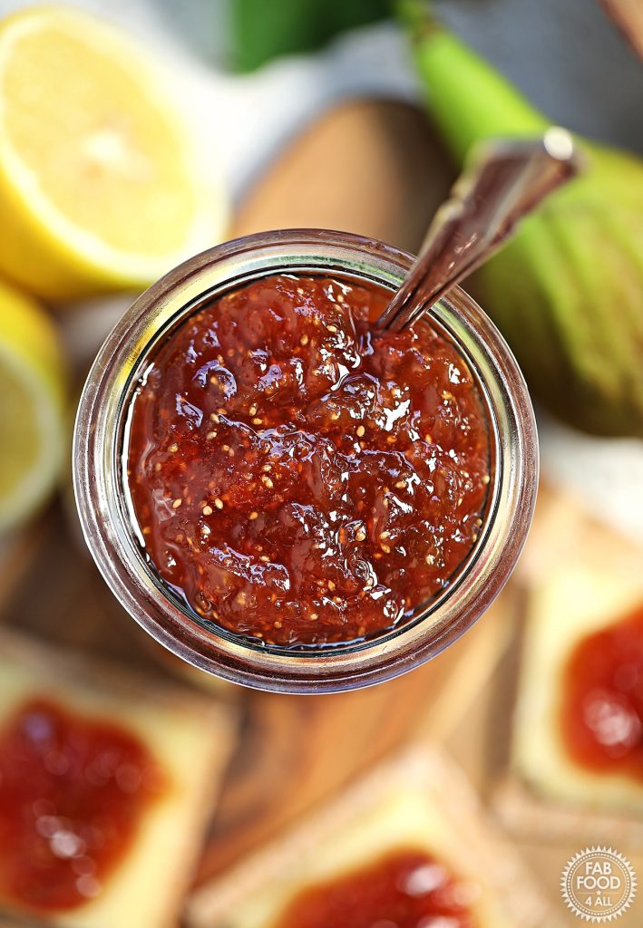 Overhead shot of open jar of Simple Fig Jam with a teaspoon resting in it. Halved lemon, fig and cheese and biscuits topped with jam blurred in the background.