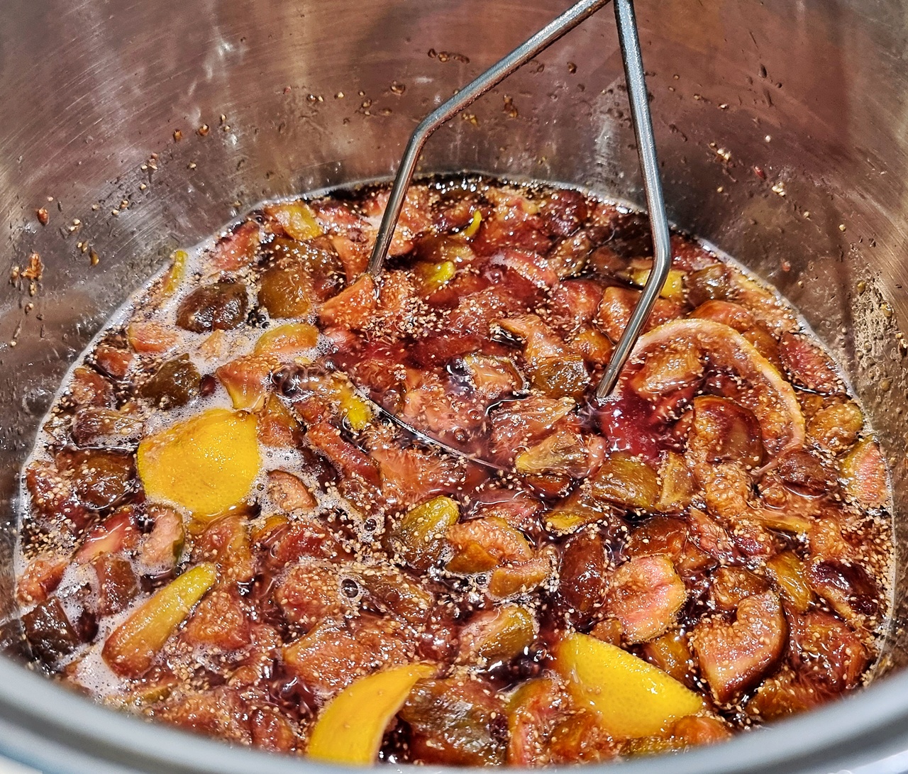 Mashing the fig jam mixture with a potato masher in a large stainless steel pot.